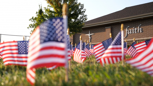 Just a few of the 2,977 flags are visible in the foreground. In the background, the Lansing Catholic cross and the word "faith" is visible on a building.
